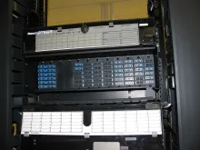 A fiber-optic patch panel mounted in a network rack, its rows of blue connectors labeled for the network rack it serves.