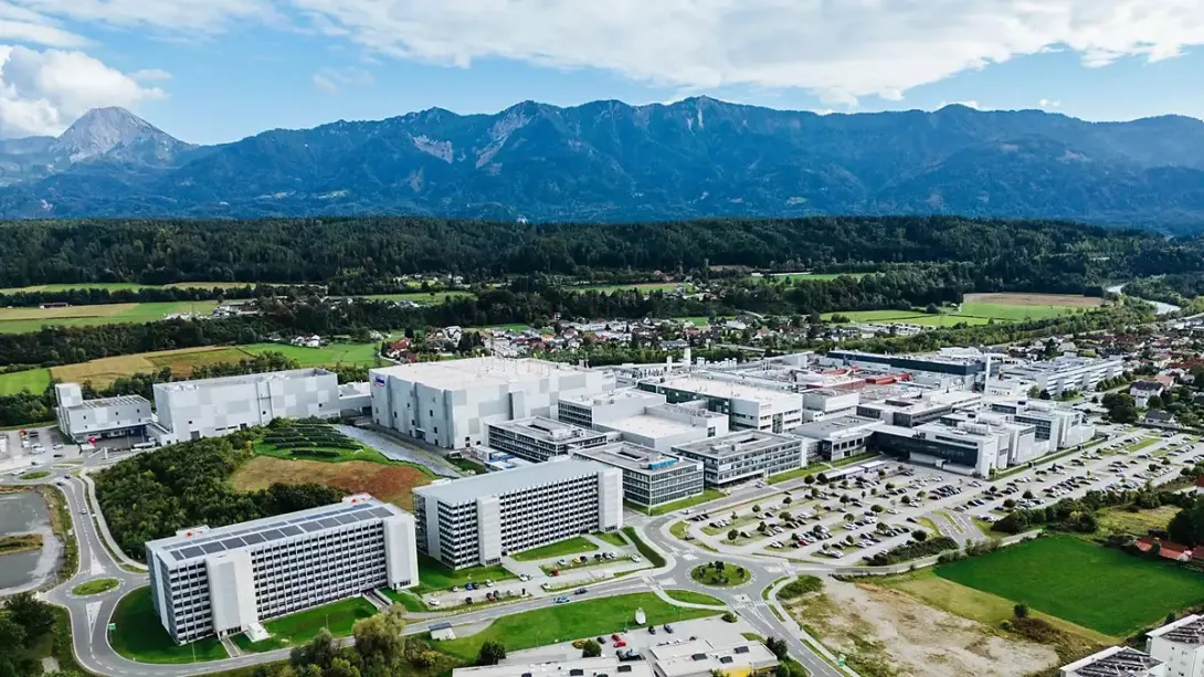 Aerial photograph of Infineon's semiconductor site in Villach, Austria, with its fab buildings set against forested hills and mountains.