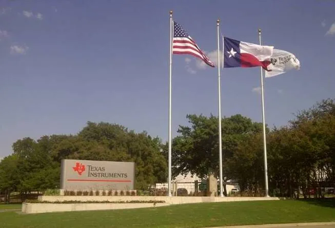 The entrance sign at Texas Instruments' North Campus facility in Dallas, Texas, with United States, Texas and company flags flying beside it.